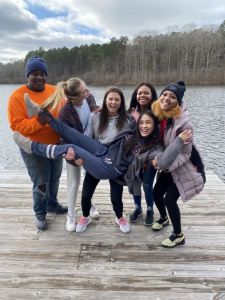 Group of individuals smiling and laughing standing on a dock holding up one person with a body of water in the backdrop